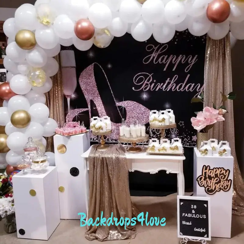 Stylish birthday party setup featuring a black and rose gold glitter backdrop with high heels and a champagne glass, surrounded by a white and gold balloon garland. The table is adorned with gold sequin fabric, desserts, party favors, and floral arrangements, creating a glamorous and festive atmosphere.
