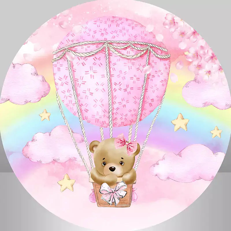A pink circular backdrop with a teddy bear in a pink hot air balloon floating in the sky with rainbow stars and flower petals
