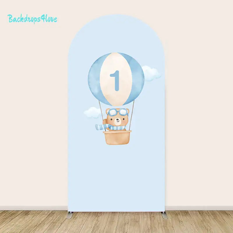 Arched backdrop featuring a teddy bear in a hot air balloon marked with the number '1,' surrounded by clouds in a pastel blue sky.